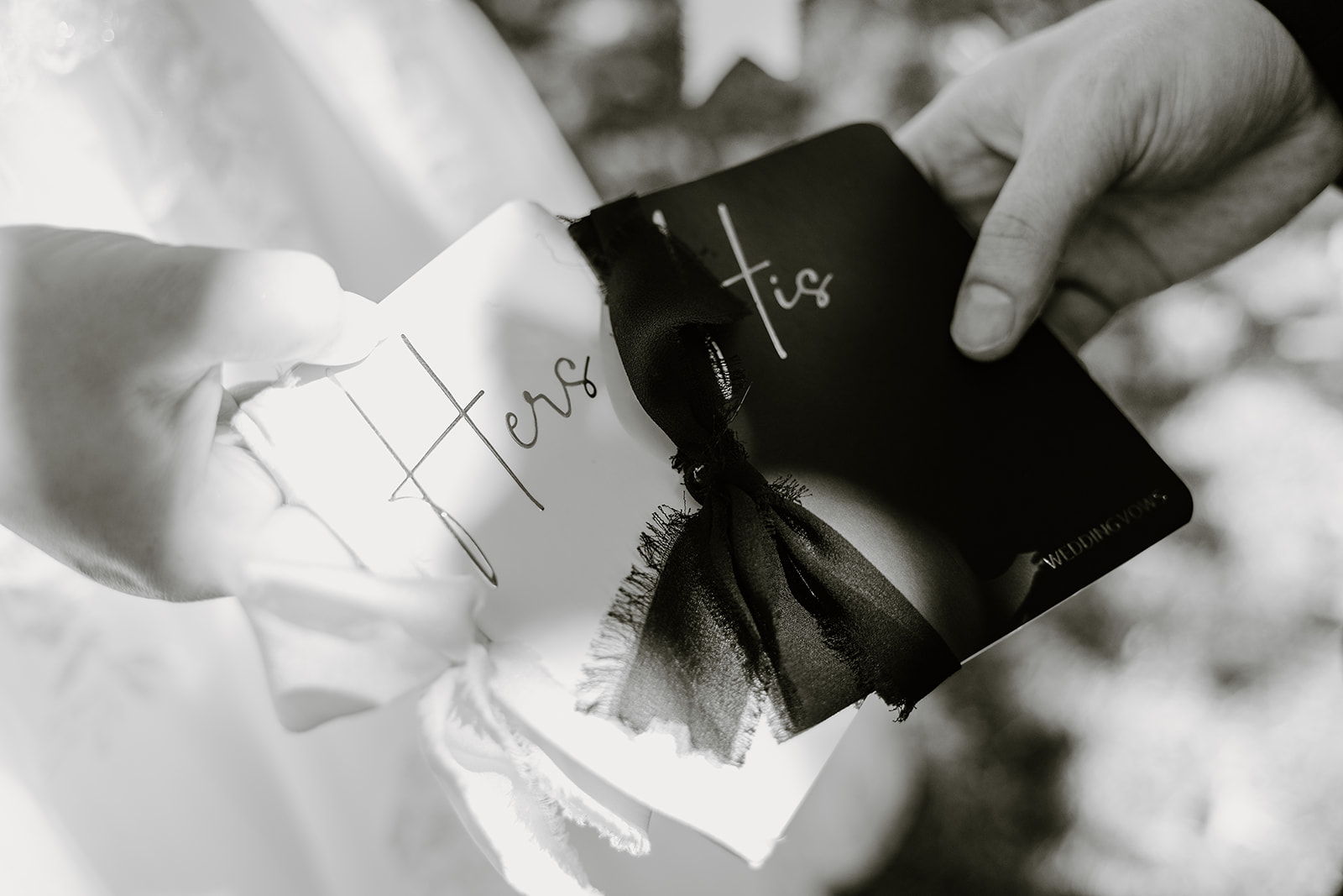 Black and white close-up of wedding vow books tied with ribbon as the couple holds them together during the ceremony.